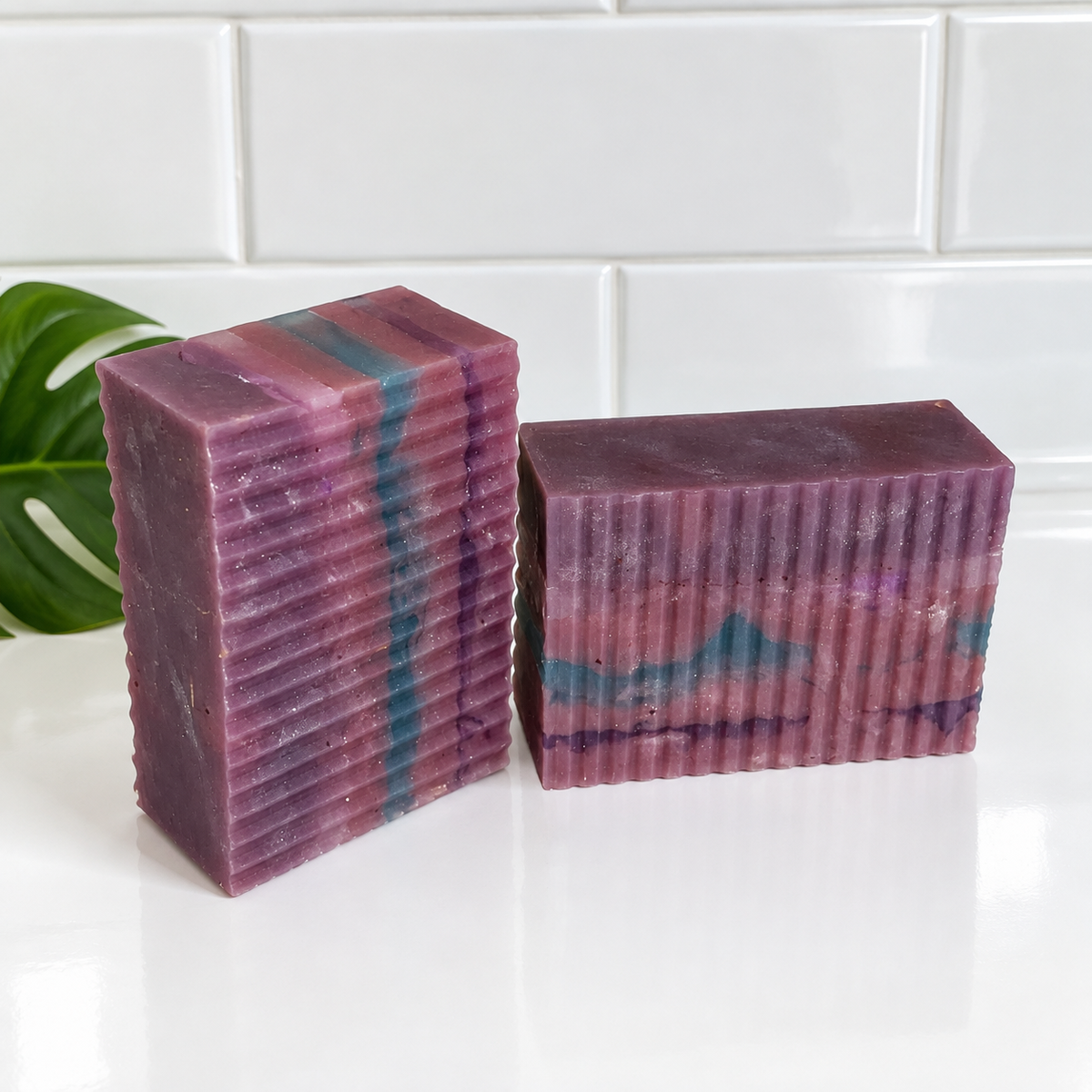 Two rectangular handmade soap bars with a ribbed texture, featuring marbled purple, pink, and teal patterns, displayed on a white surface with a white subway tile background.