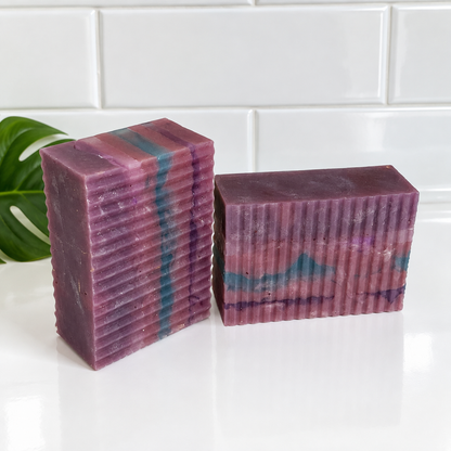 Two rectangular handmade soap bars with a ribbed texture, featuring marbled purple, pink, and teal patterns, displayed on a white surface with a white subway tile background.