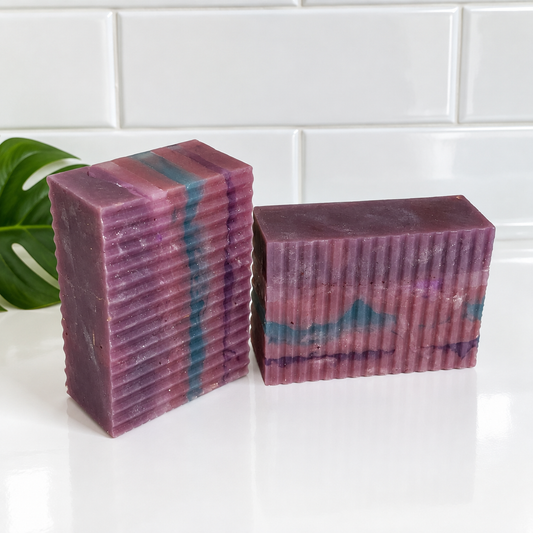 Two rectangular handmade soap bars with a ribbed texture, featuring marbled purple, pink, and teal patterns, displayed on a white surface with a white subway tile background.