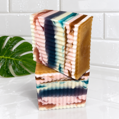 Two stacked rectangular bars of layered handmade soap with horizontal stripes of brown, pink, cream, teal, and dark blue, set on a white surface with a green leaf in the background.