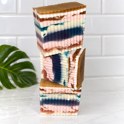 Three stacked rectangular bars of layered handmade soap on a white marble surface with a white subway tile backsplash. A green monstera leaf is visible on the left.