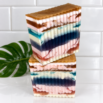 Two stacked rectangular bars of layered handmade soap with a wavy ribbed edge, featuring pink, blue, white, and brown horizontal layers, set against a white tiled background with a green leaf prop.