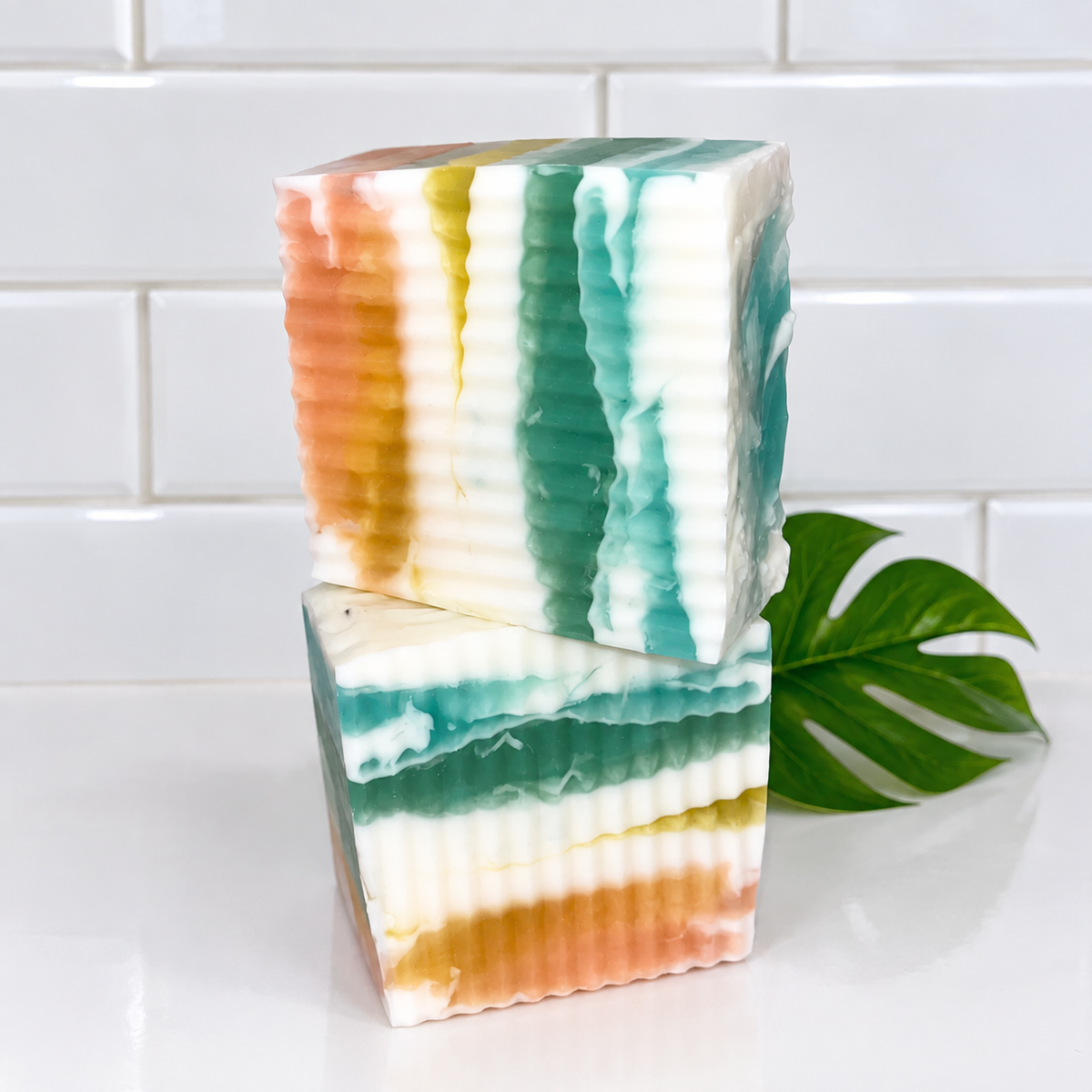 Two stacked rectangular bars of marbled handmade soap with vertical ridges, featuring stripes of teal, white, peach, and yellow, set against a white subway tile background with a green monstera leaf prop.