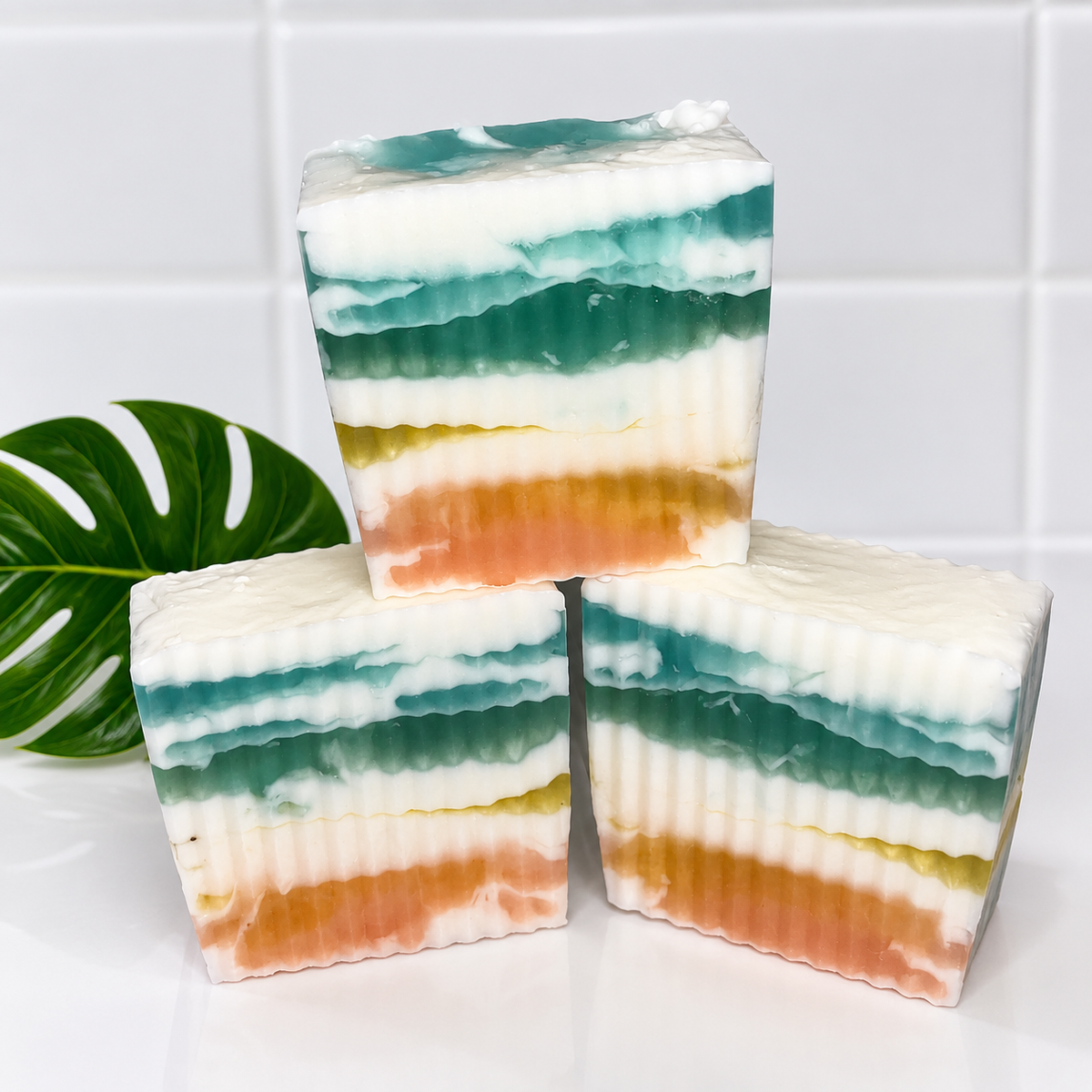Three layered rectangular bars of soap stacked in a pyramid arrangement on a white surface, with a green monstera leaf prop on the left. The soap has horizontal layers of white, teal, green, yellow, and peach-orange.