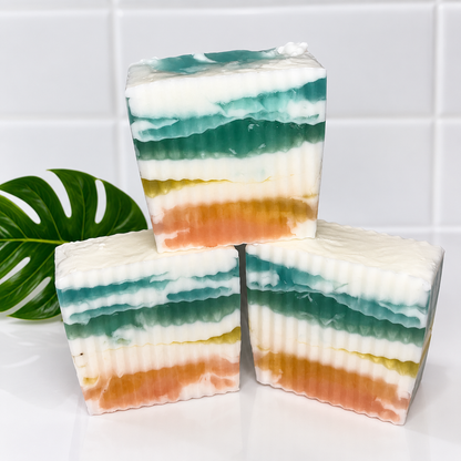 Three layered rectangular bars of soap stacked in a pyramid arrangement on a white surface, with a green monstera leaf prop on the left. The soap has horizontal layers of white, teal, green, yellow, and peach-orange.