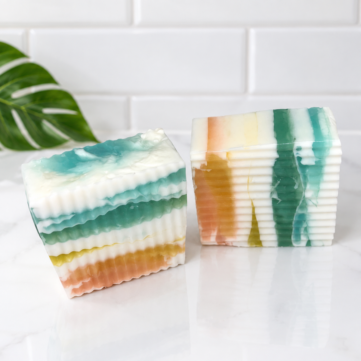 Two rectangular handmade soaps with a multicolor layered design, one in teal, white, and orange, and the other in green, white, and orange, displayed on a white marble surface with a white subway tile background and a green leaf prop.