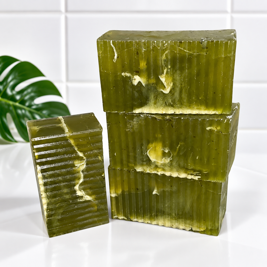 Three rectangular bars of translucent olive green soap with a ribbed texture, stacked on a white surface with a white tiled background and a green leaf prop on the left.