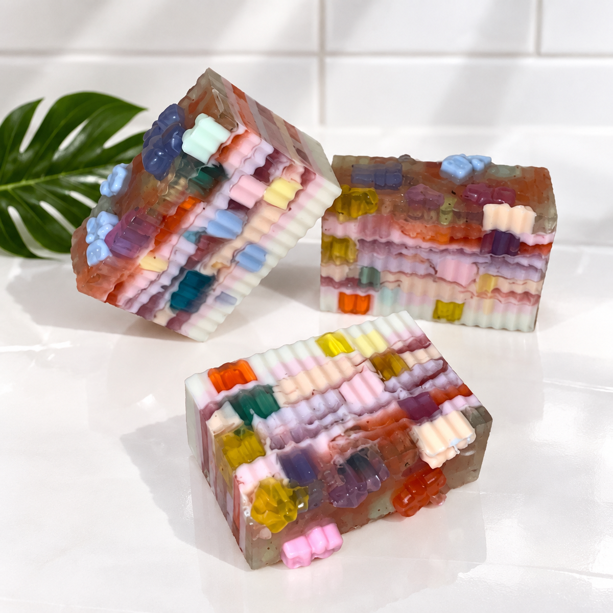 Three handmade layered soap bars decorated with colorful gummy bears, displayed on a glossy white surface with a green leaf in the background.