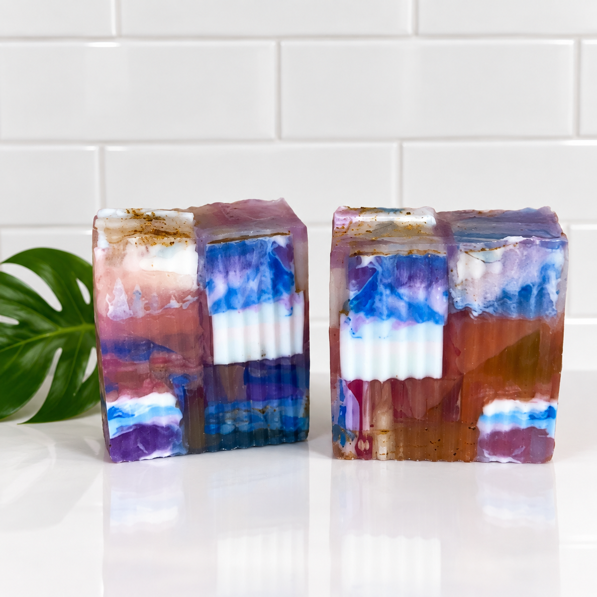 Two handmade layered soap bars with a marbled pattern of blue, purple, white, and reddish-brown, displayed on a white surface with a green leaf in the background.