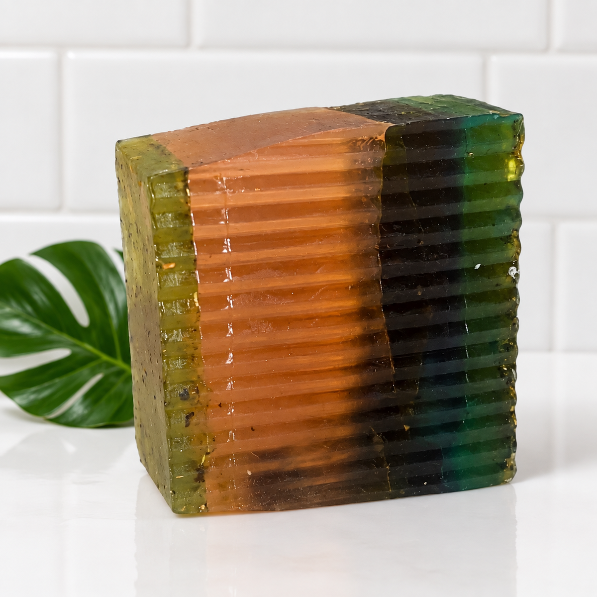 A handmade rectangular bar of soap with a ribbed texture, featuring a tri-color gradient of orange, dark green, and black, styled on a white surface with a green monstera leaf and white subway tiles in the background.