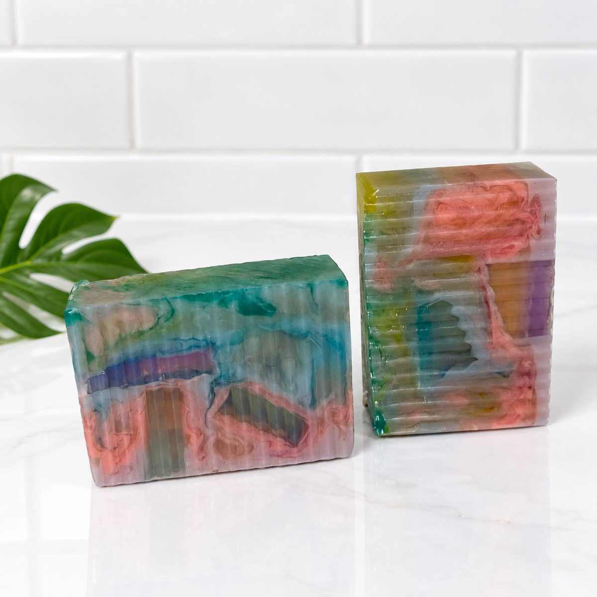 Two handmade marbled soaps in vibrant green, pink, and purple tones, displayed on a white marble surface with a white subway tile background and a green leaf prop.