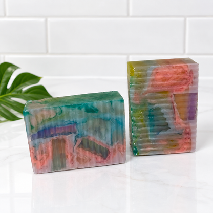 Two handmade marbled soaps in vibrant green, pink, and purple tones, displayed on a white marble surface with a white subway tile background and a green leaf prop.