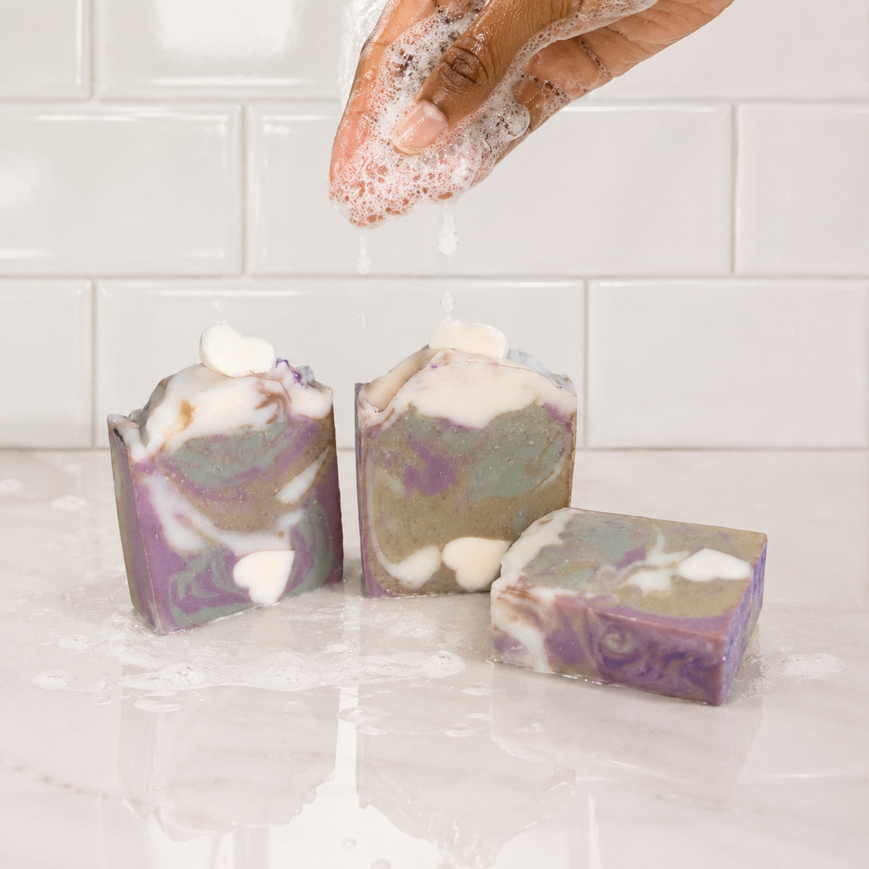Three bars of soap with a hand rubbing soap suds above them on a tiled bathroom floor.
