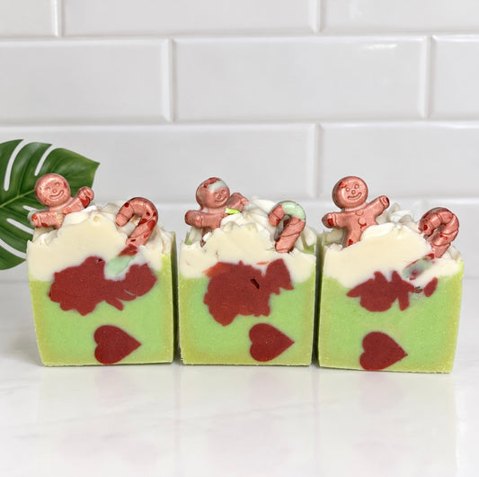 Three rectangular handmade soap bars with a light green base, white top layer, dark red heart and abstract shapes, and gingerbread men decorations, arranged on a white surface against a white subway tile background.