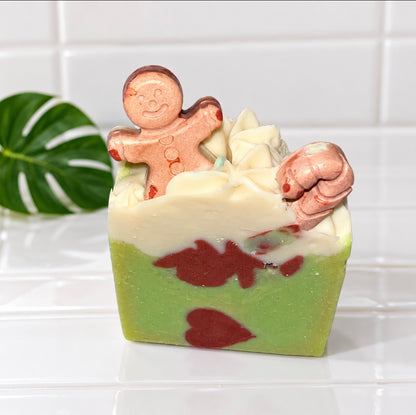 A decorative layered soap bar with a light green base, a thick white middle layer, and dark red marbled accents, topped with a gingerbread person figure and a pinkish-brown ornament, set on a white tiled surface with a green leaf in the background.
