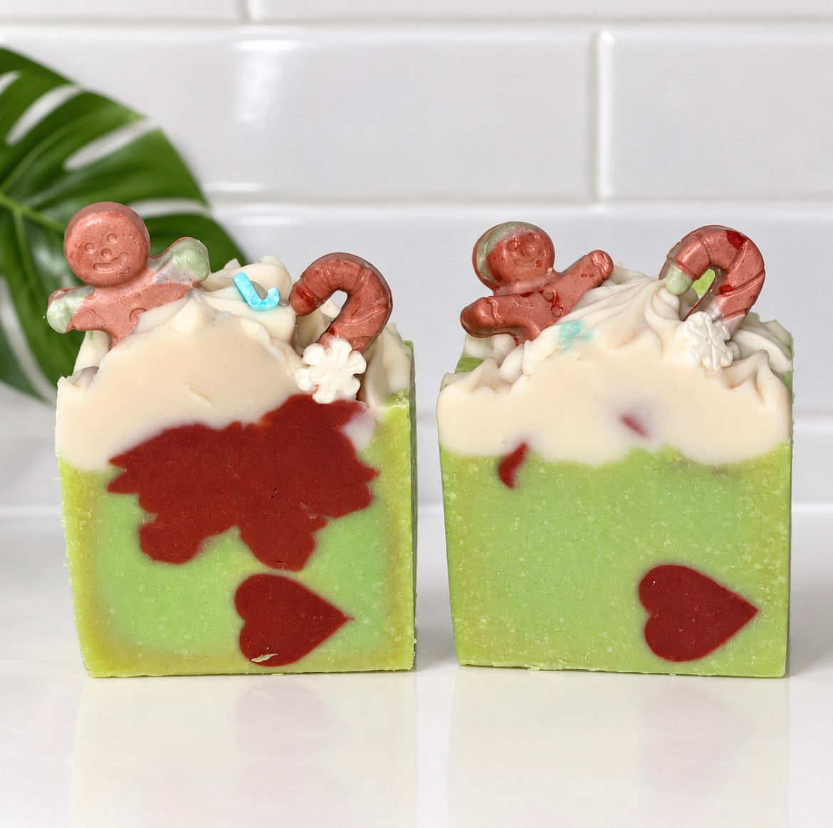 Two decorative handmade soap bars with gingerbread-themed embellishments, featuring a light green base, white cream top, and red heart and candy cane details, set against a white tiled background.