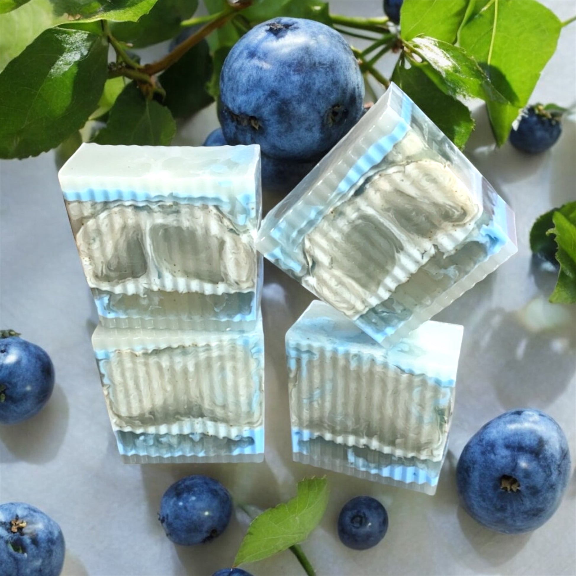 Clear soap bars with blueberry slices on a white surface with blueberries and leaves.