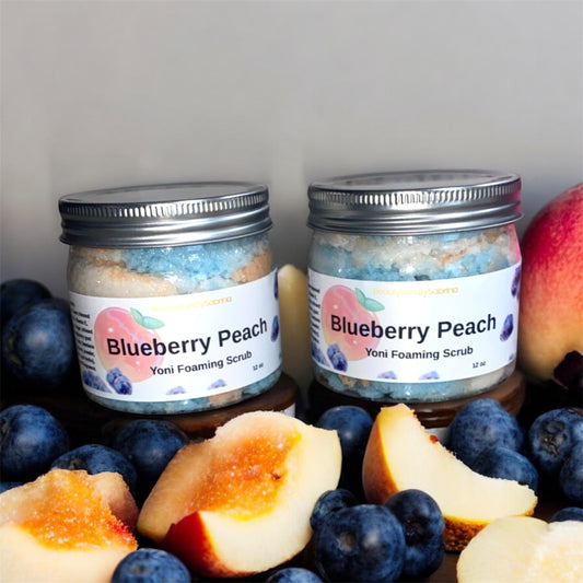 Two jars of Blueberry Peach Yoni Foaming Scrub surrounded by blueberries, peaches, and figs on a gray background.