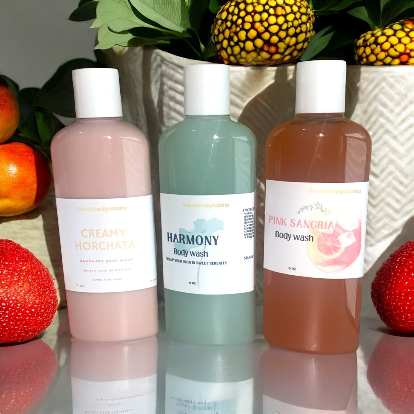 Three bottles of body wash with labels on a reflective surface with decorative fruits and plants in the background.